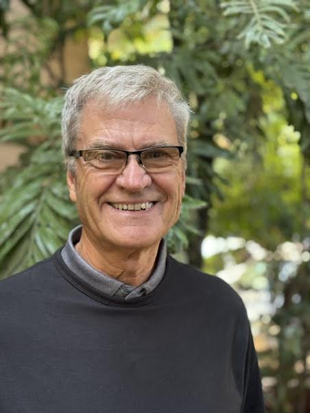 Smiling portrait of Curtis Coughlan, a Kenya Relief Board member wearing glasses and a dark grey polo overtop a green collared shirt against lush green foliage on campus in Migori.